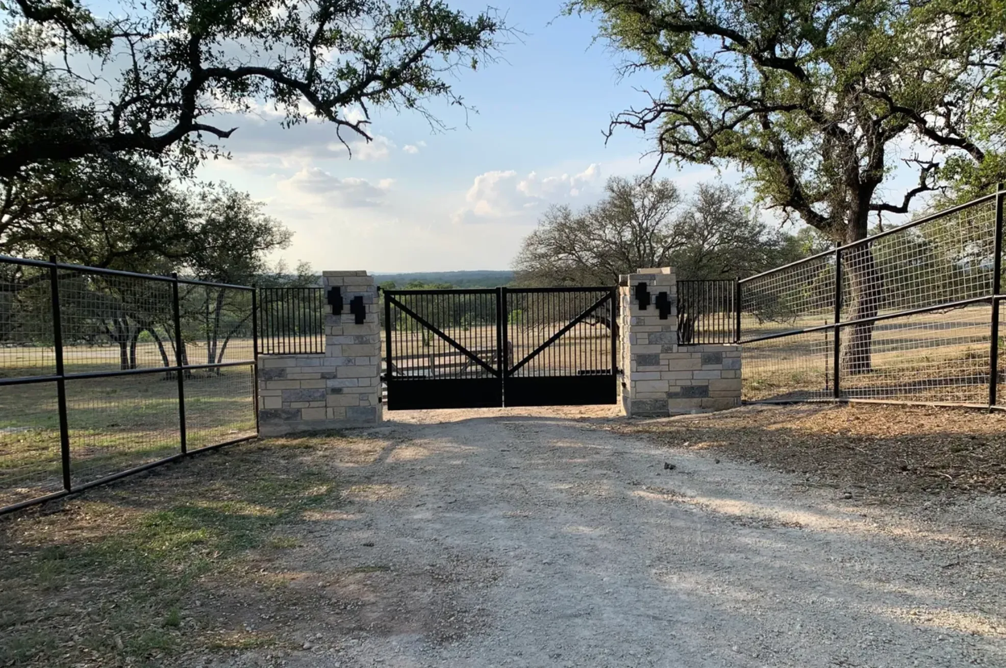 Custom black steel ranch gate and fencing between limestone pillars under live oaks