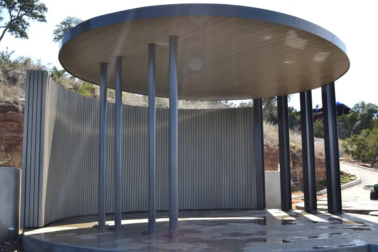 Round steel pavilion with slender columns, a slatted circular roof and a curved metal wall