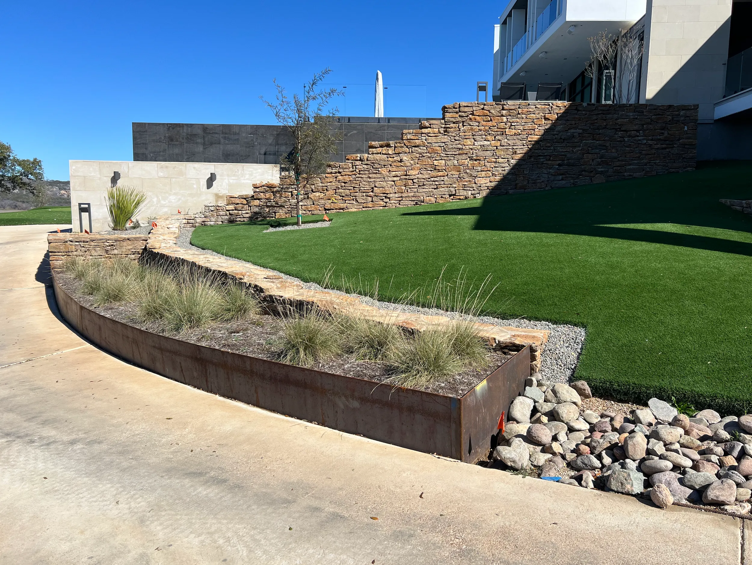 Curving weathering-steel landscape edging along a driveway below a stone retaining wall
