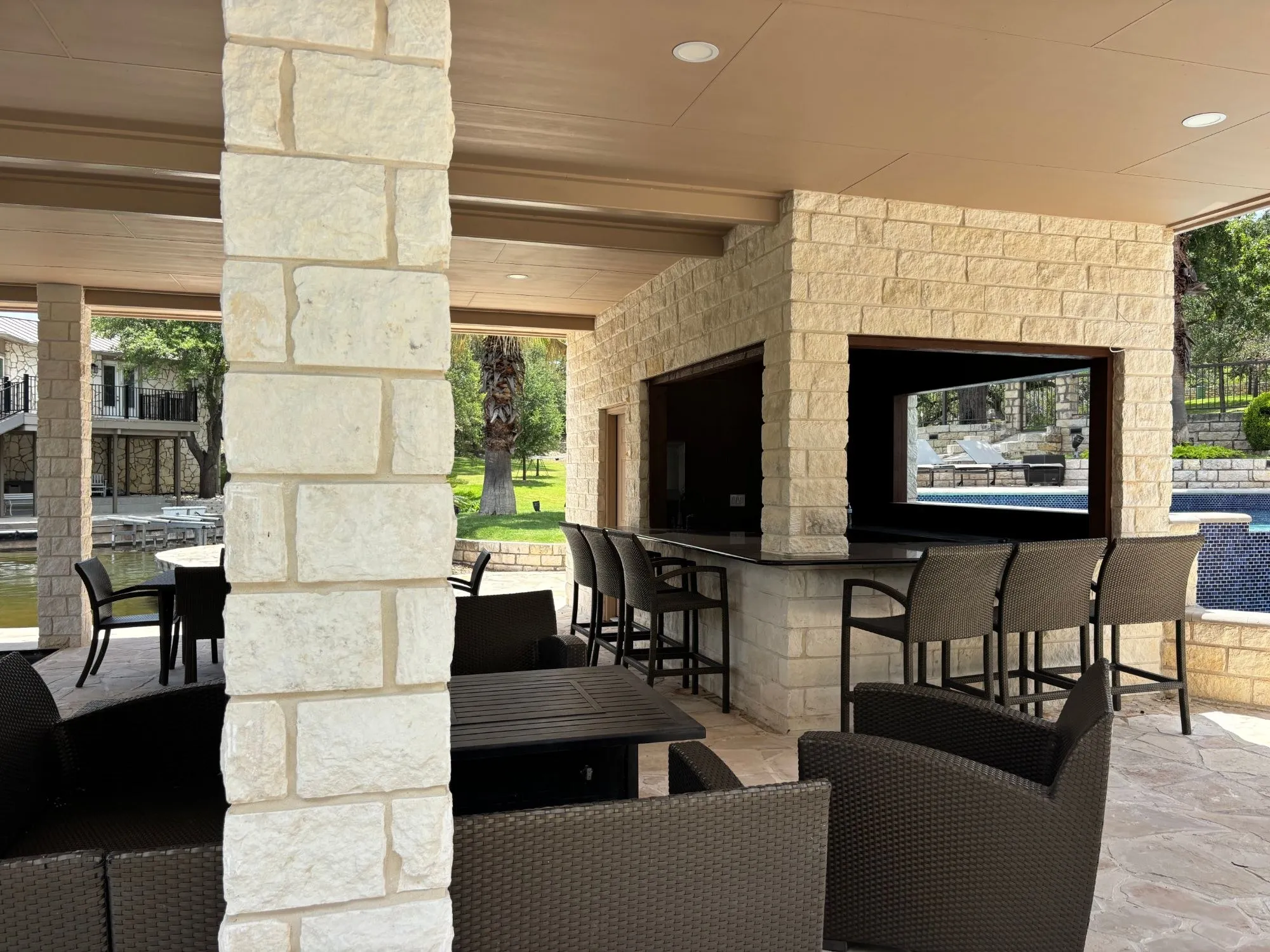 Stone outdoor bar with a dark countertop and bar stools on two sides, with recessed lights in the ceiling above