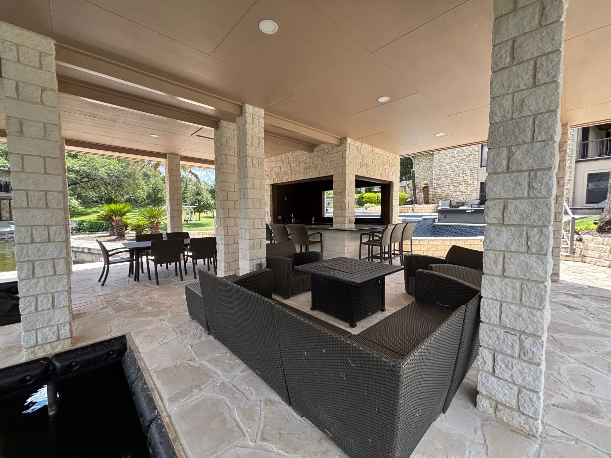 Wide view under the pavilion roof across the lounge, dining table and stone bar toward the pool and house